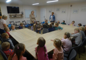 Grupa dzieci siedzi przy stolikach i spogląda na uśmiechniętą do nich panią. Po lewej stronie stoi pani Agnieszka a po prawej stronie uśmiechnięta pani dyrektor.